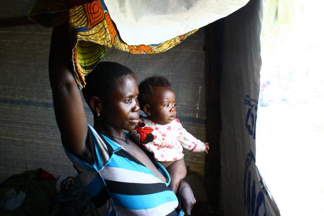 Marie regarde par la fenêtre découpée dans les bâches en plastique de l’abri collectif dans lequel elle vit avec sa famille. Restera-t-elle ici ou retournera-t-elle bientôt dans l’est de la RDC ? Dieu seul le sait, dit-elle. Photo : Grace Cahill/Oxfam Marie dans son abri en plastique